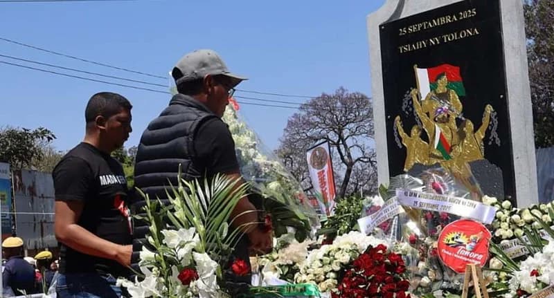 Two men lay floral tributes at a black memorial monument in Madagascar, commemorating a political struggle. The plaque features an illustration of people raising the national flag and bears the inscription 'TSIAHY NY TOLONA' under the date September 25, 2025.
