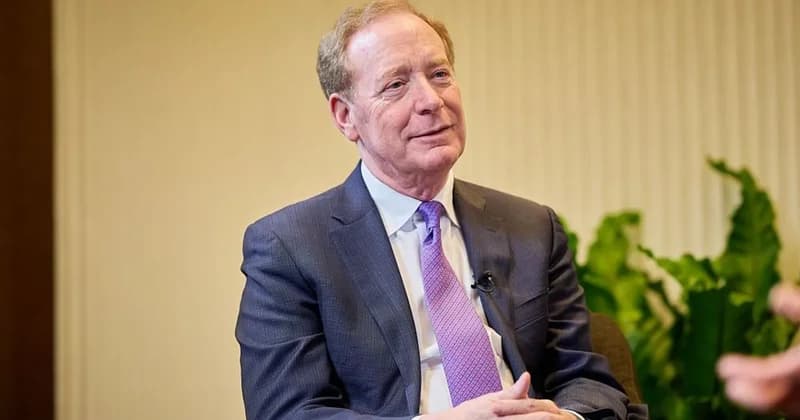 Brad Smith, the President of Microsoft, is pictured in a formal interview setting wearing a dark suit and a patterned purple tie. He appears to be listening or speaking during a discussion, with a lapel microphone visible on his jacket and green foliage in the background.