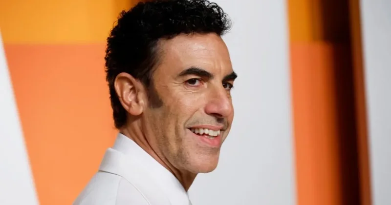 A close-up portrait of British actor Sacha Baron Cohen smiling against a blurred background of orange and white panels. He is wearing a white garment, likely a suit jacket or dress shirt, in what appears to be a red-carpet or promotional event setting.