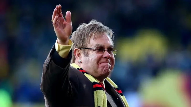 Sir Elton John is seen waving to a crowd while wearing a yellow and black Watford FC scarf. He has a somber or pained facial expression, and the background shows a blurred view of a stadium crowd.