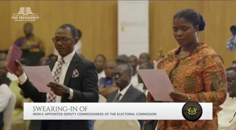 A man in a suit and a woman in a patterned traditional dress are seen during a formal swearing-in ceremony. Both individuals are holding documents and reciting their oaths, with the man holding up a Bible as they are inducted as Deputy Commissioners of the Electoral Commission of Ghana.