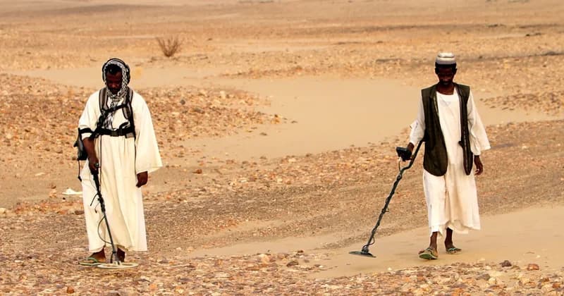 Two men in traditional attire traverse a vast, arid desert landscape while using handheld metal detectors to scan the rocky ground. They appear to be engaged in artisanal gold prospecting, a common but dangerous activity in border regions like the one between Egypt and Sudan.