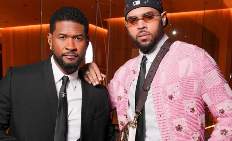 American R&B singers Usher and Chris Brown pose together for a photograph. Usher is dressed in a formal black suit with a white shirt and black tie, while Chris Brown wears a pink patterned cardigan, a black cap, and tinted glasses.