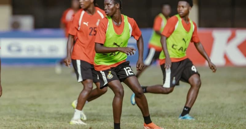 Players from the Ghana women's national football team, known as the Black Queens, are captured during a focused training session on a grass pitch. They are wearing orange jerseys, lime green training bibs, and black shorts featuring the Ghana Football Association crest.