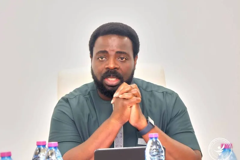 A portrait of Dr. Da-Costa Aboagye, the Chief Executive of the National Health Insurance Authority (NHIA), speaking at an event with his hands clasped. Several water bottles and a tablet are on the table before him, and the NHIS logo is watermarked in the bottom right corner.
