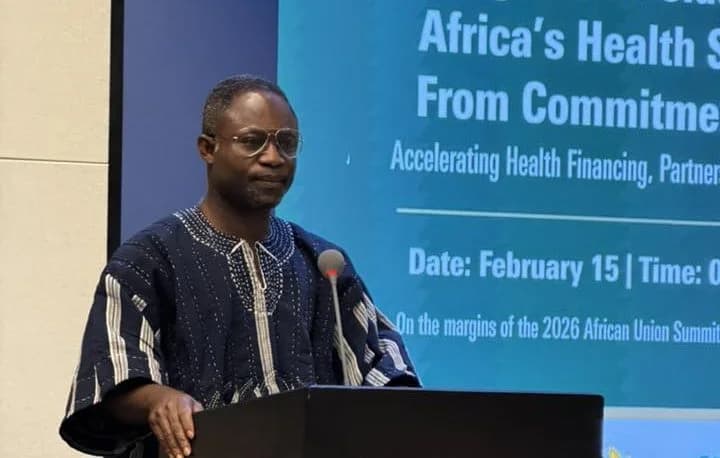 Hon. Kwabena Mintah Akandoh, Ranking Member on the Health Committee of Parliament, is pictured speaking at a podium. He is wearing a traditional blue and white striped fugu and eyeglasses while addressing an audience at a health-related summit on the margins of an African Union event.