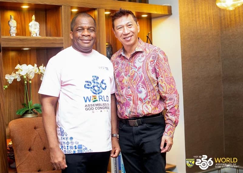 Two religious leaders smile while standing together indoors, ahead of the World Assemblies of God Congress in Ghana. The man on the left wears a white t-shirt branded for the event, while the man on the right wears a vibrant, patterned shirt.