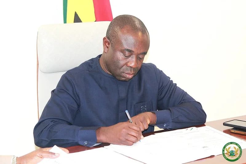 Ghanaian Minister for Employment and Labour Relations, Ignatius Baffour-Awuah, is seen signing an official agreement at a desk. The photograph captures the moment of signing against the backdrop of the national flag, representing a formal government ceremony related to overseas job creation.