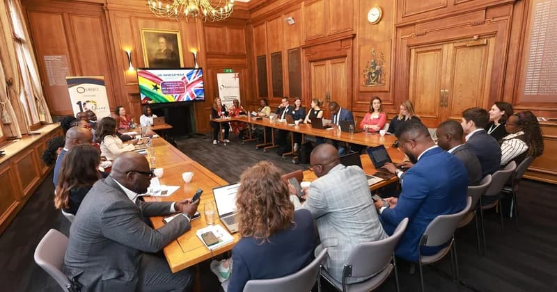 A wide-angle photograph showing a group of business professionals and delegates seated around a large U-shaped wooden table in a classic wood-paneled conference room in London. The background features a digital display for the 'UK Investment Roadshow' flanked by the Ghana and UK flags, along with banners for the UK-Ghana Chamber of Commerce (UKGCC).