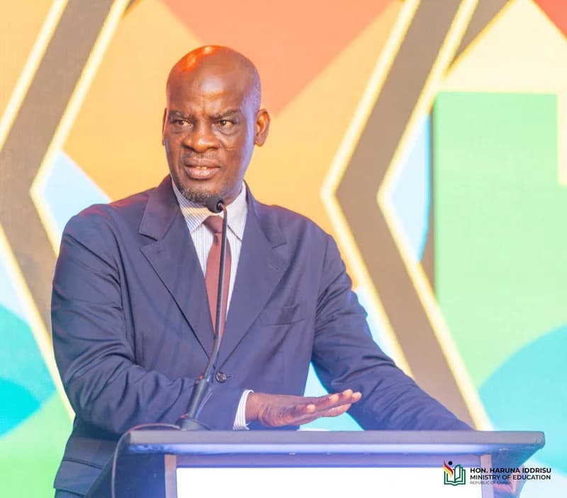 Hon. Haruna Iddrisu, Member of Parliament for Tamale South, is captured speaking at a formal engagement while standing behind a podium. He is dressed in a dark navy suit and reddish-brown tie, set against a vibrant geometric background with a Ministry of Education logo visible.