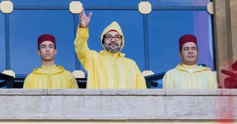 King Mohammed VI of Morocco waves from a balcony flanked by two men, including Crown Prince Moulay El Hassan, all wearing traditional yellow djellabas. The scene captures the monarch in a formal public appearance, consistent with state ceremonies or national addresses in Morocco.