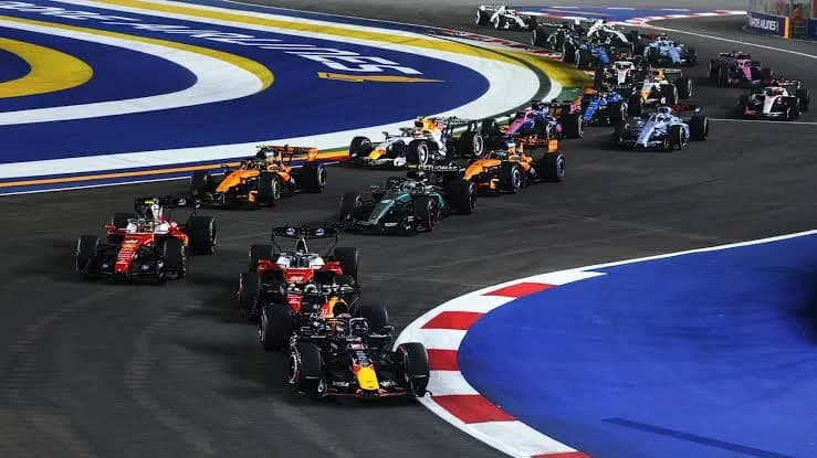 A large group of Formula 1 cars navigate a sharp corner on a lighted street circuit during a night race. The pack includes prominent teams such as Red Bull Racing, Ferrari, and Mercedes-AMG, showcasing the high-intensity competition characteristic of the sport.