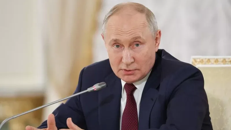 A close-up photograph of Russian President Vladimir Putin speaking during a formal meeting. He is wearing a navy blue suit and a maroon tie, positioned behind a microphone with a formal background featuring gold-trimmed furniture.