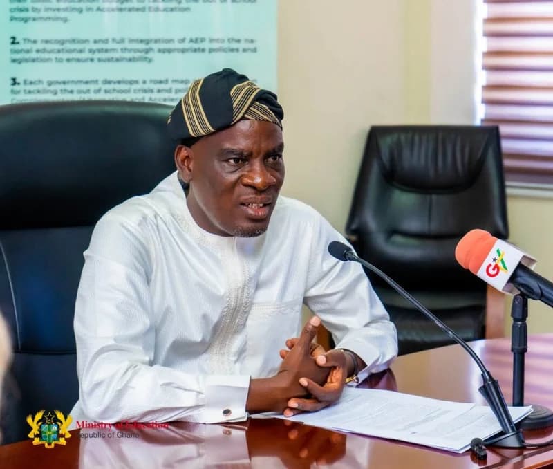 Dr. Yaw Osei Adutwum, Ghana's Minister for Education, is shown speaking during an official meeting. He is dressed in a white traditional outfit and a striped fugu cap, seated at a desk with a GTV microphone and documents in front of him.