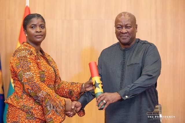 President John Mahama is pictured presenting a scroll adorned with the colors of the Ghana flag to a woman during a formal ceremony. Both individuals are shaking hands in front of a wood-paneled wall, indicating an official government appointment or state function.
