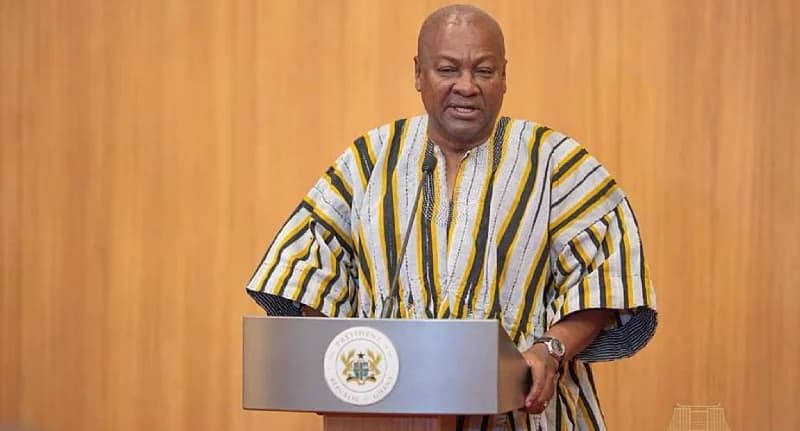 President John Dramani Mahama of Ghana is captured delivering a speech behind a presidential podium. He is wearing a traditional striped smock while addressing an audience against a wooden paneled background.
