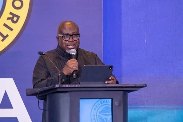 An official from the Ghana Revenue Authority (GRA) addresses the media or public from behind a podium. He is speaking into a microphone while referring to a tablet, set against a blue backdrop featuring the GRA logo.