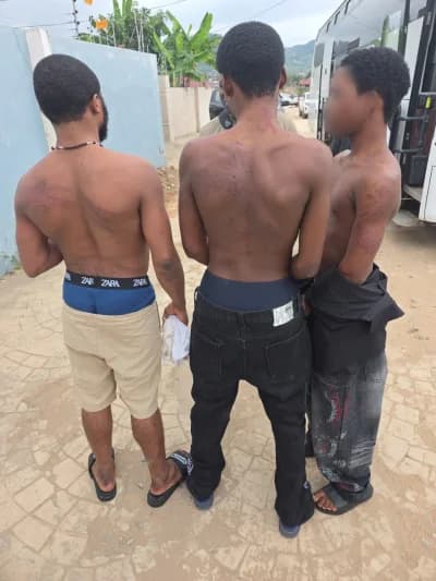 Three shirtless young men are shown from behind, apparently in police custody following a cybercrime crackdown in Ghana. The individuals exhibit visible marks on their backs and are standing in an outdoor area next to a white vehicle.