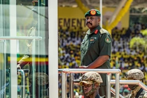 General Muhoozi Kainerugaba of the Ugandan People's Defence Force stands at a public event, dressed in a green military uniform and beret. He is positioned behind a railing with soldiers in camouflage gear in the foreground and a large crowd in yellow attire visible in the background.