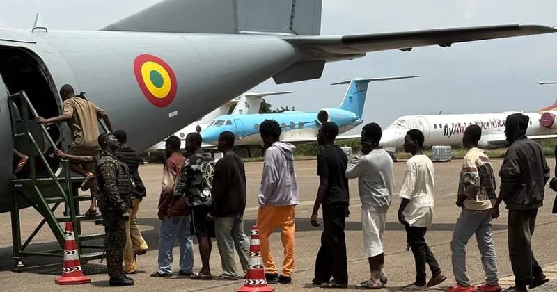 A group of young men are seen boarding a grey Ghana Air Force transport plane on a tarmac under the supervision of a soldier in camouflage. The aircraft prominently displays the Ghanaian national roundel, with other commercial and private aircraft visible in the background.