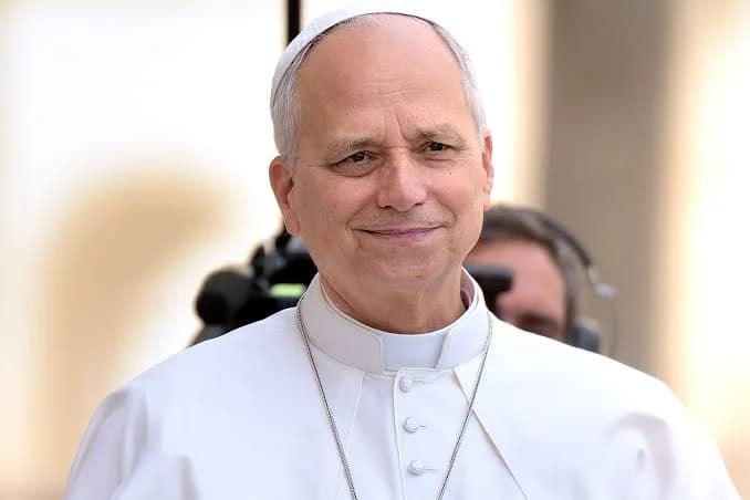 A medium close-up photograph of a man identified as Pope Leo, wearing traditional white papal vestments and a zucchetto. He has a calm expression with a slight smile, positioned against a blurred background that includes media equipment and another person wearing headphones.