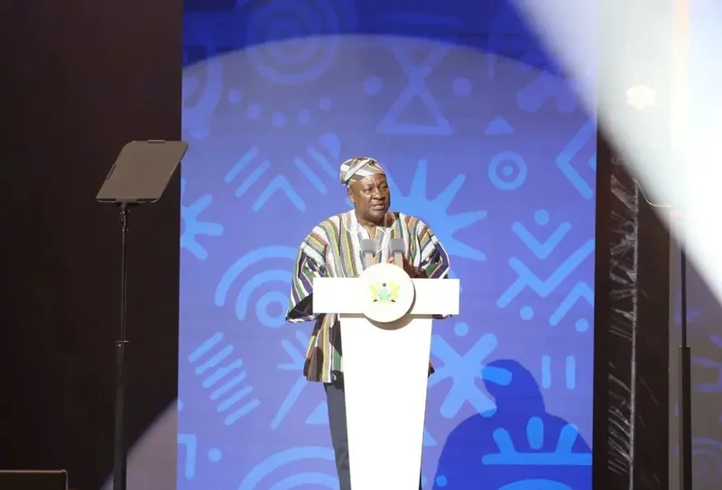 President John Mahama is seen delivering a speech from a white podium adorned with the Coat of Arms of Ghana. He is dressed in a traditional multicolored smock (batakari) and matching cap, standing against a vibrant blue backdrop featuring Adinkra-style patterns.