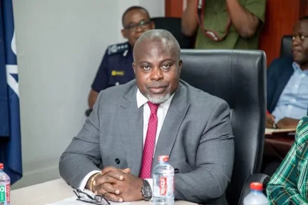 A photograph of a high-ranking official, likely from the Ghana Revenue Authority (GRA), seated at a formal meeting or press briefing. He is wearing a grey suit and a pinkish-red patterned tie with his hands clasped on a table that holds water bottles and his eyeglasses. In the background, other individuals are visible, including a police officer in uniform and a person holding a professional camera.