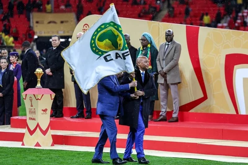 CAF President Patrice Motsepe and an official are seen carrying the Confederation of African Football (CAF) flag across a football pitch during a formal ceremony. The background features a red and gold podium, the AFCON trophy on a stand, and several officials in formal attire within a stadium setting.