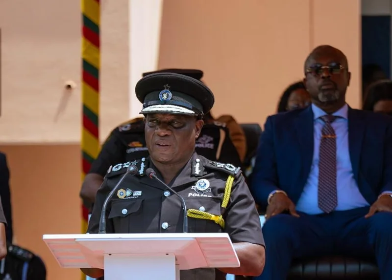A high-ranking Ghana Police Service official, likely Inspector General of Police Dr. George Akuffo Dampare, is pictured speaking at a white lectern. He is dressed in a formal black police uniform adorned with several medals and insignia, while other officials and the Ghanaian national colors are visible in the background.