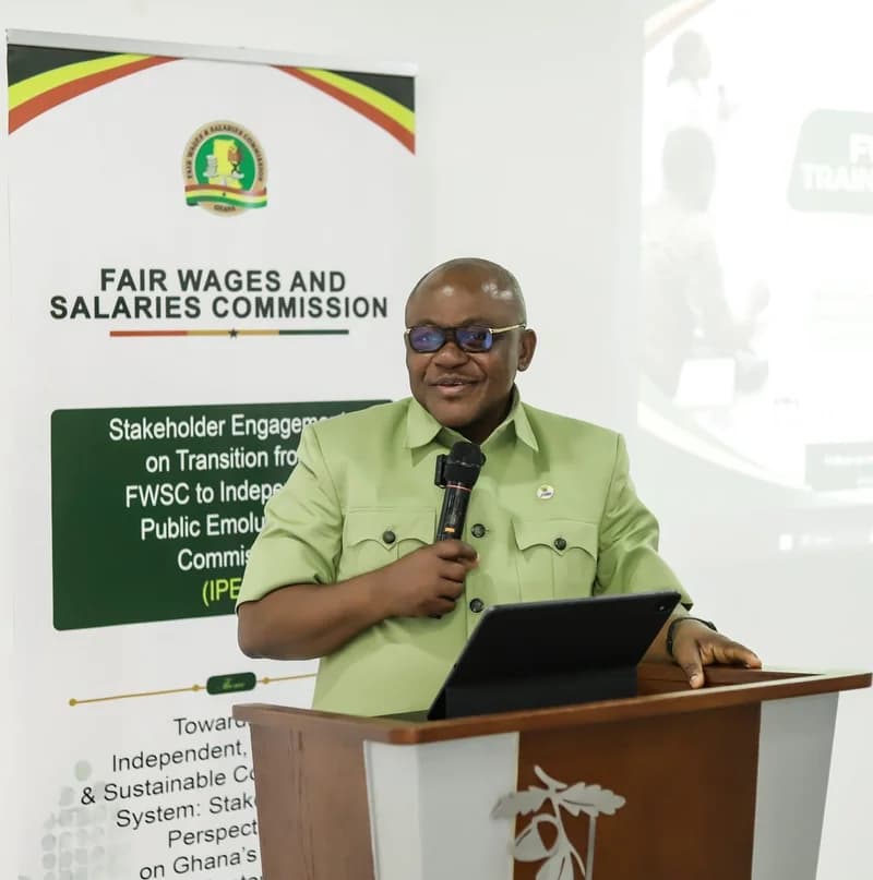 Ben Arthur, the Chief Executive of the Fair Wages and Salaries Commission (FWSC), is pictured speaking at a stakeholder engagement session. He stands at a wooden podium with a tablet, positioned beside a banner detailing the transition to the Independent Public Emoluments Commission (IPEC).