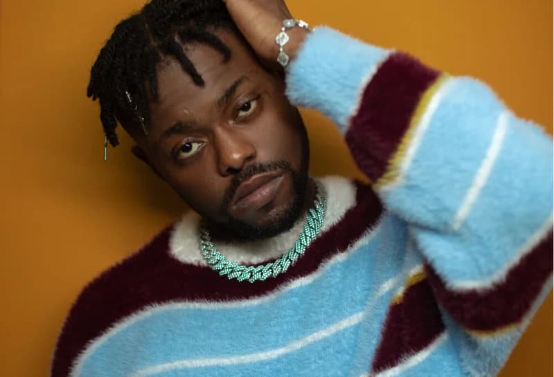 A close-up portrait of the Nigerian musician Lojay, wearing a textured, multi-colored striped fuzzy sweater in blue, maroon, and white. He is posed with one hand resting on his head against a solid mustard yellow background, looking directly into the camera lens with a serious expression.