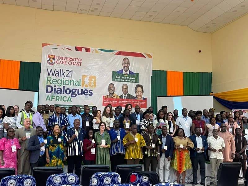 A large group of delegates pose for a photograph at the Walk21 Regional Dialogues Africa conference held at the University of Cape Coast. The participants are gathered in front of a large event banner that features the University of Cape Coast logo and photos of key speakers including the Vice-Chancellor and the Ghanaian Minister for Transport.