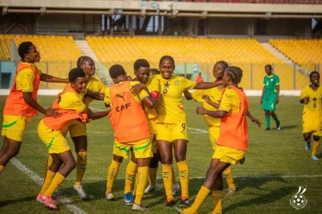 A group of Ghana's Black Maidens football players are seen celebrating joyfully on a football pitch. They are wearing their yellow national kits and orange training bibs, with a large stadium containing empty yellow seats visible in the background.