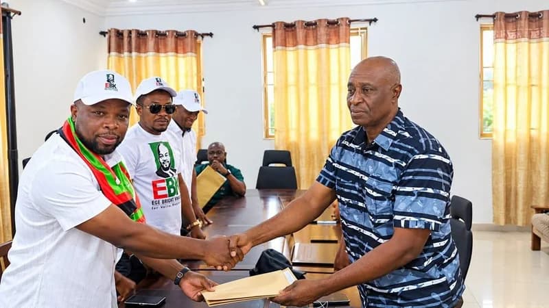 A photograph showing two men shaking hands across a meeting table while exchanging a brown envelope. The man on the left wears a sash in the National Democratic Congress (NDC) colors, while supporters in the background wear 'EBK' branded white t-shirts and caps.