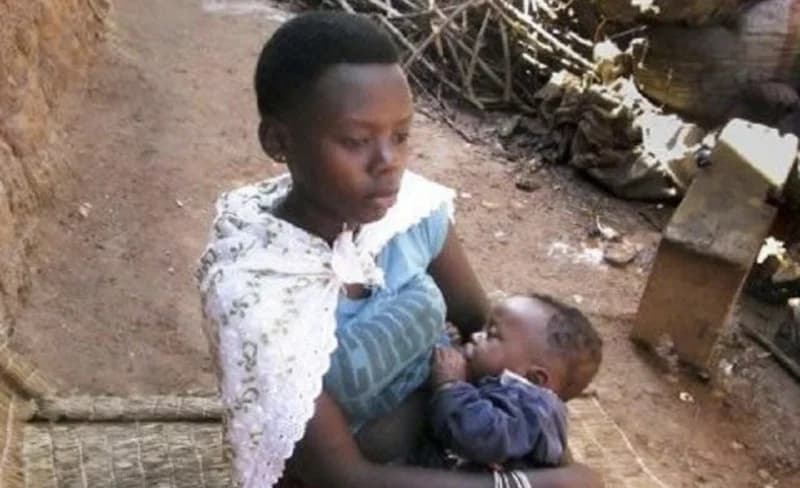 A young African woman, appearing to be a teenager, sits outdoors while breastfeeding an infant. The setting features a dirt ground and rustic environment, reflecting a rural or low-income urban context often associated with discussions on social health.