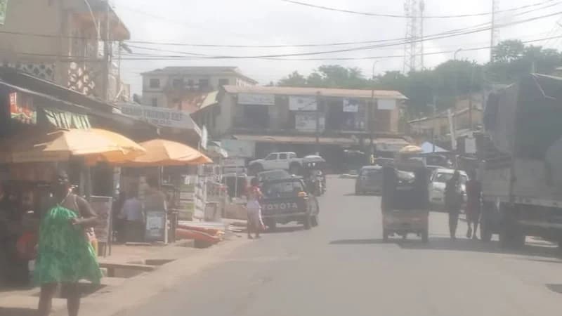 A street view in Cape Coast, Ghana, depicting daily economic activity with local storefronts, street vendors under orange umbrellas, and various vehicles including a Toyota pickup truck. The scene shows the bustling urban environment and typical trading infrastructure found in the region.