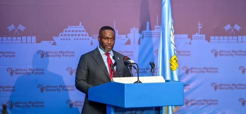A man in a formal dark suit and red tie delivers a speech at a blue podium during the African Maritime Security Forum. The background features a thematic blue wall with silhouettes of ships and the forum's logo, accompanied by a light blue flag on the right.