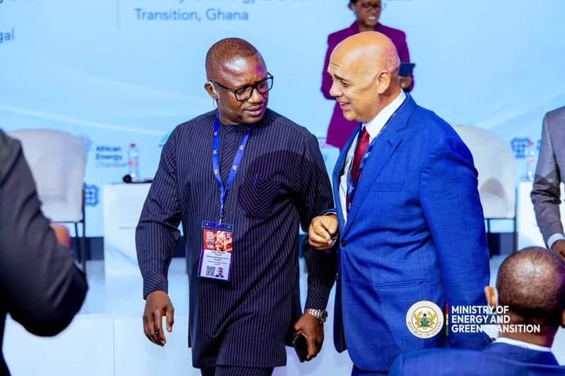 A photograph of two officials engaged in conversation during an energy conference in Ghana. One man is wearing a dark, striped traditional garment with a VIP lanyard, while the other is in a blue business suit. The event branding in the background includes the Ministry of Energy and Green Transition logo.