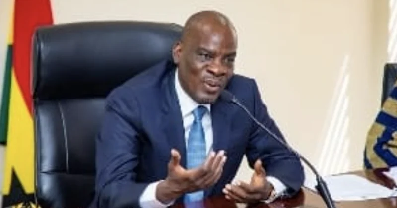 Haruna Iddrisu, a prominent Ghanaian politician, is captured speaking into a microphone during an official engagement. He is dressed in a dark blue suit and seated in front of a Ghanaian flag, indicating a formal government or parliamentary context.