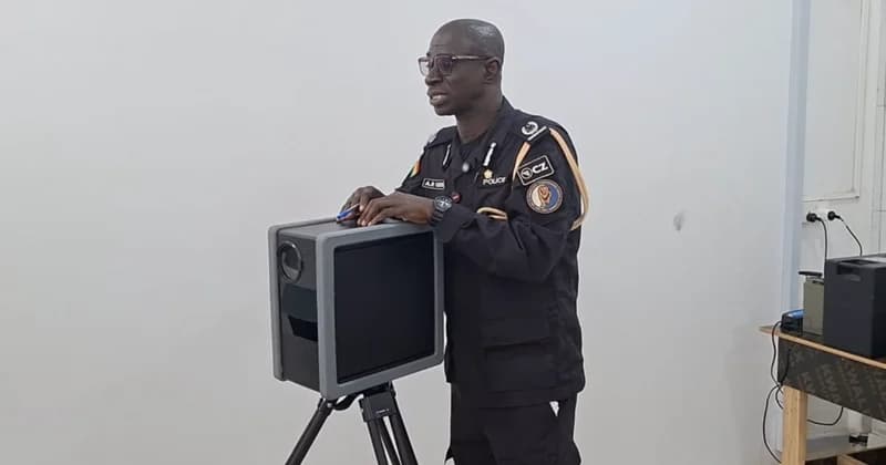 A high-ranking Ghana Police Service officer stands next to an automated traffic enforcement camera mounted on a tripod. He is dressed in a black service uniform and appears to be demonstrating or explaining the TraffiTech-GH technology used for road safety monitoring.