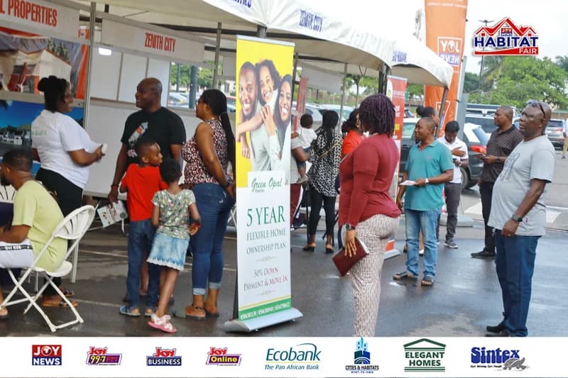 A photograph showing the JoyNews Habitat Fair event with attendees interacting at various exhibition booths under white canopies. A prominent roll-up banner for Green Opal Properties displays a 5-year flexible home ownership plan, while several corporate sponsor logos appear at the bottom.