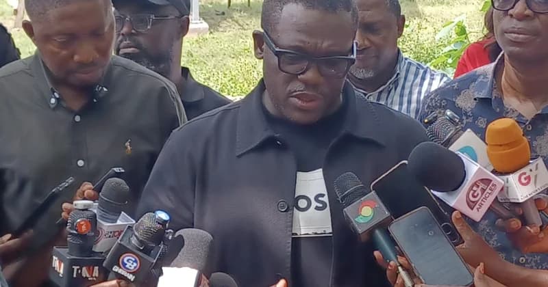 Ghanaian Member of Parliament Sam Nartey George is captured addressing a press briefing. He is surrounded by multiple microphones from various media organizations, indicating a public announcement or interview regarding national policy.