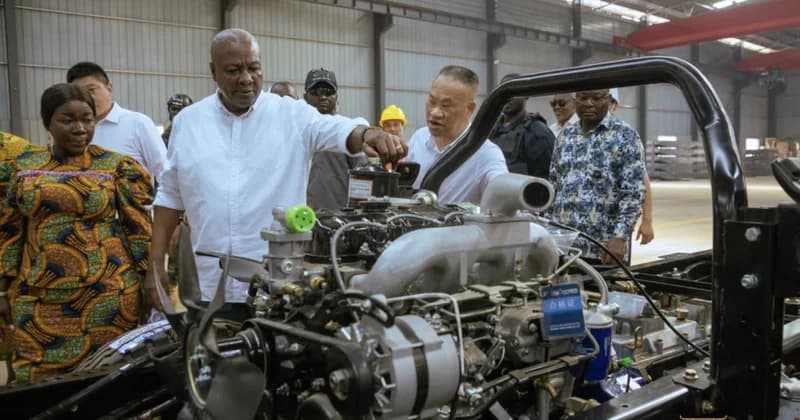 President John Dramani Mahama is seen inspecting a vehicle engine during a high-profile visit to an industrial facility in the Afienya/Tema area. He is accompanied by several officials and factory managers who are briefing him on the manufacturing processes and technical specifications of the equipment.
