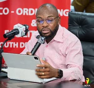 A photograph of Pius Enam Hadzide, the CEO of the National Youth Authority (NYA), speaking into a microphone while referencing a tablet during a press briefing. He is seated at a desk against a red backdrop that displays official agency text, and the microphone features 'LUV FM' branding.