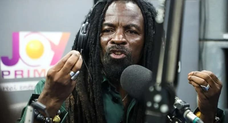 A close-up photograph of Ghanaian musician Rocky Dawuni during a studio interview. He is wearing headphones and speaking passionately into a microphone, with the Joy Prime logo visible in the blurred background.
