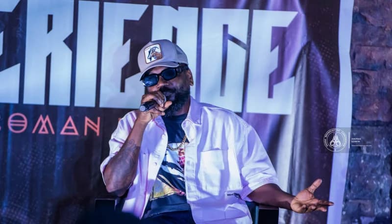 Ghanaian musician Samini is captured speaking into a microphone during a public event or interview. He is dressed in a white shirt, a graphic t-shirt, and a grey cap with a goat patch, wearing sunglasses as he gestures with his left hand.