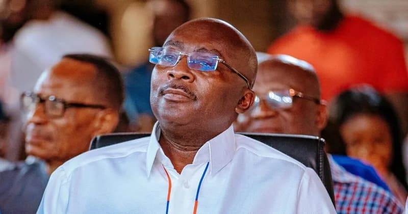 Dr. Mahamudu Bawumia is captured in a close-up shot during a formal event. He is wearing rimless glasses and a crisp white shirt while looking attentively forward, with other blurred figures visible in the background.