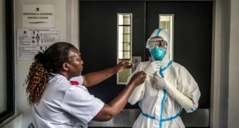 A medical professional is shown assisting a colleague with the donning of full personal protective equipment (PPE), including a hooded hazmat suit, turquoise mask, and goggles. They are standing in a clinical hallway next to a sign detailing instructions for donning and doffing equipment, likely in preparation for treating patients with infectious diseases.