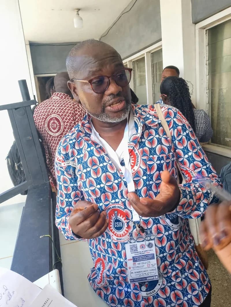 A photograph of Dr. Gordon Akon-Yamga speaking at the National Scientific Conference. He is wearing a patterned shirt with the Council for Scientific and Industrial Research (CSIR) logo and an identification badge while gesturing with his hands during an interview or discussion.