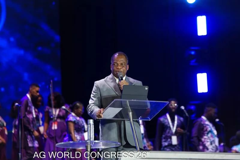 Reverend Stephen Wengam is pictured delivering a speech at the Assemblies of God (AG) World Congress. He is dressed in a grey suit and stands behind a transparent podium equipped with a tablet and microphone, with several people visible in the background on a brightly lit stage.